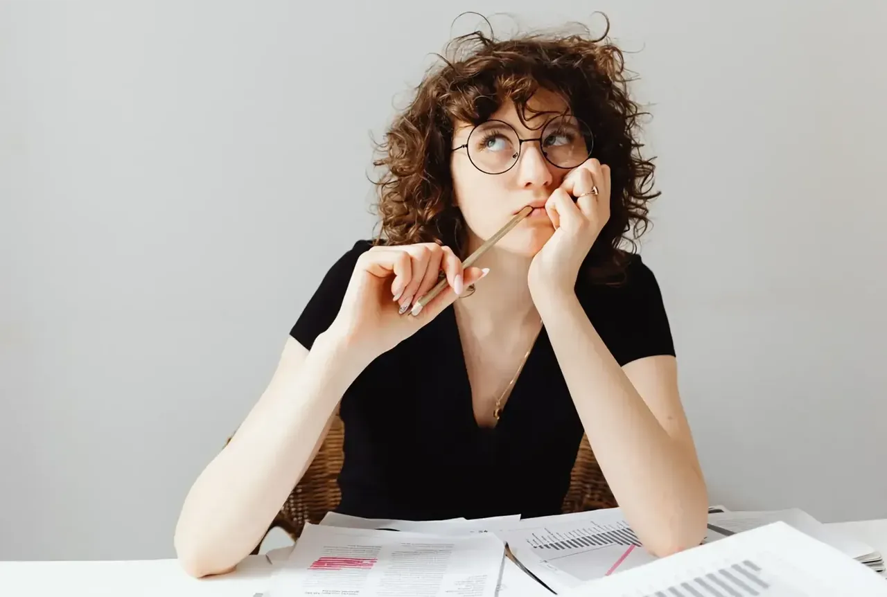 Eine junge Frau sitzt an einem Schreibtisch vor einem Stapel Papierblätter mit Berechnungen, Diagrammen und Texten und denkt nach, während sie in die Ferne blickt.